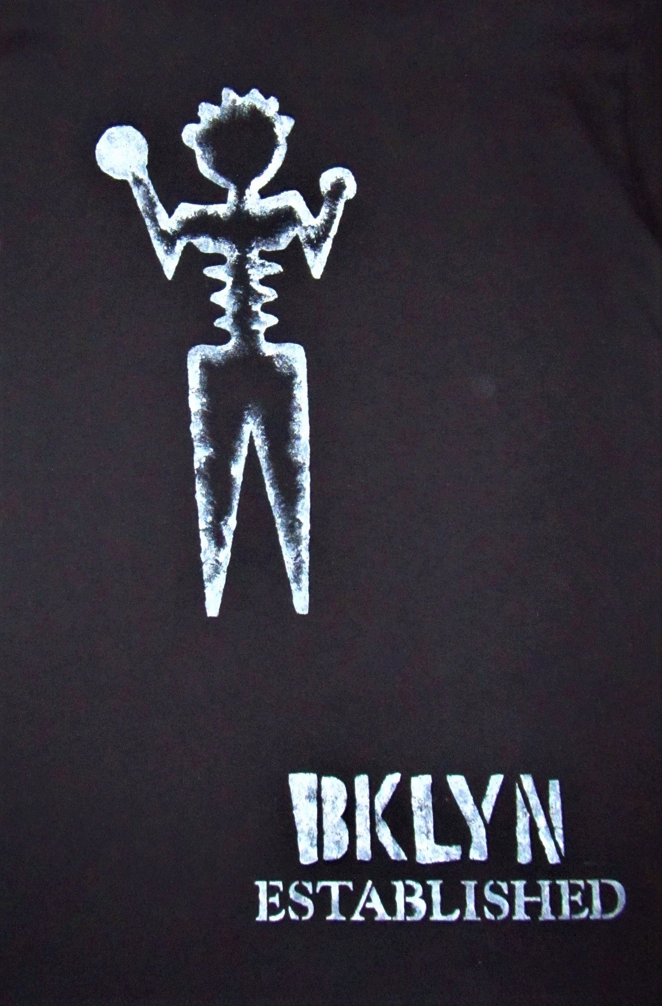 Close up of white hand-painted ambiguous person with Brooklyn established words on black tee shirt.