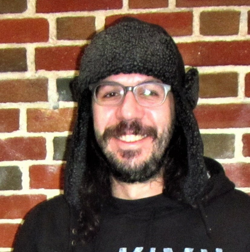 A man wearing the black and grey hooded Crowned Shelter hat with a furry brim and ear flaps.